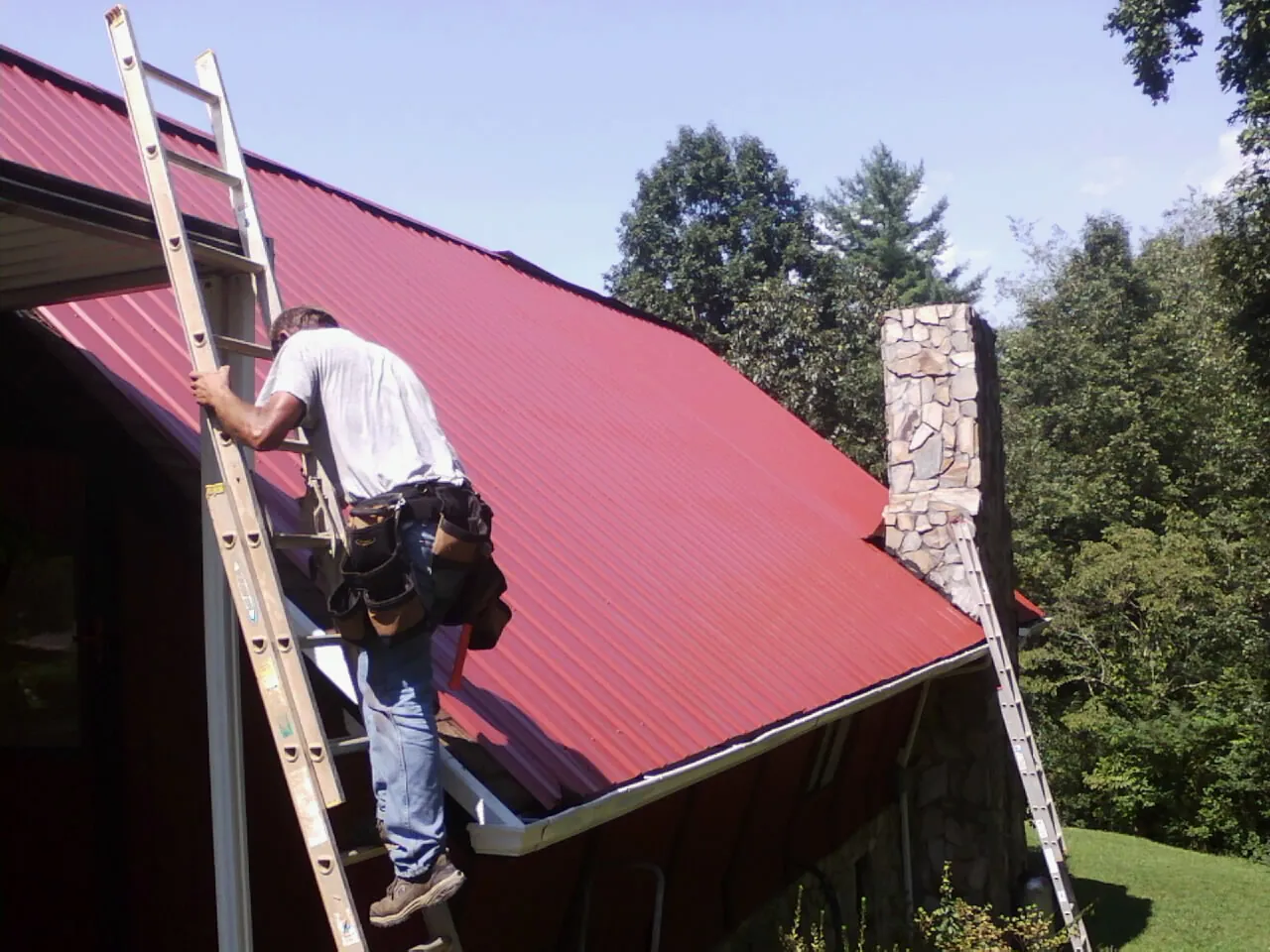 Roofer on a ladder beside a red metal roof and stone chimney — Soffit & Fascia Repair by Roofing Brothers in Mountain Top